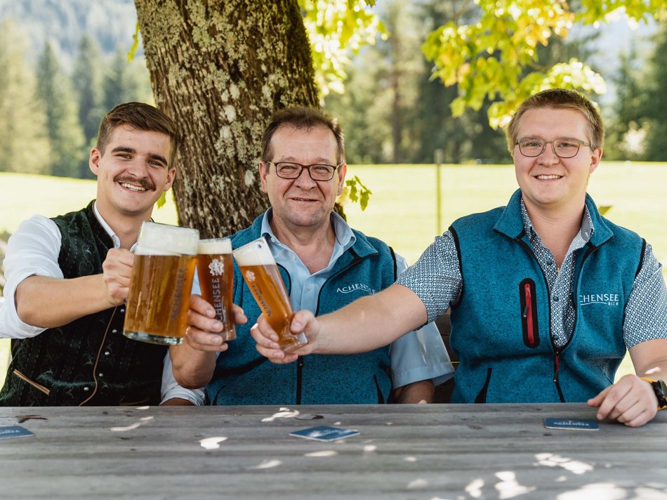 The Karlwirt’s Achenseebier Three men cheers with beer glasses outdoors under a tree