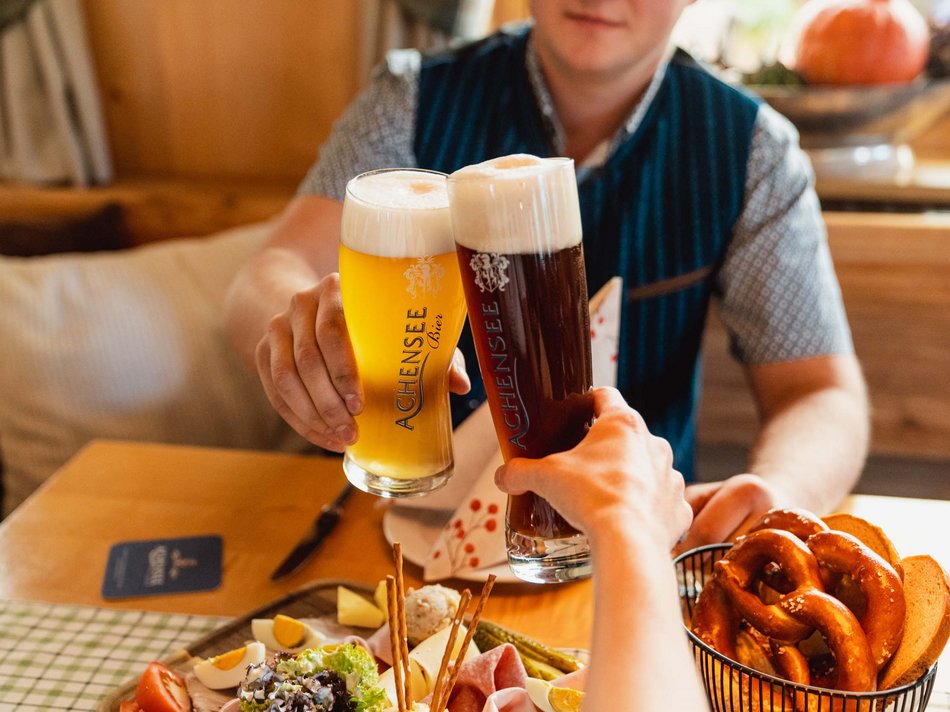 The Karlwirt’s Achenseebier People clinking glasses of light and dark Achensee beer with traditional snacks including pretzels