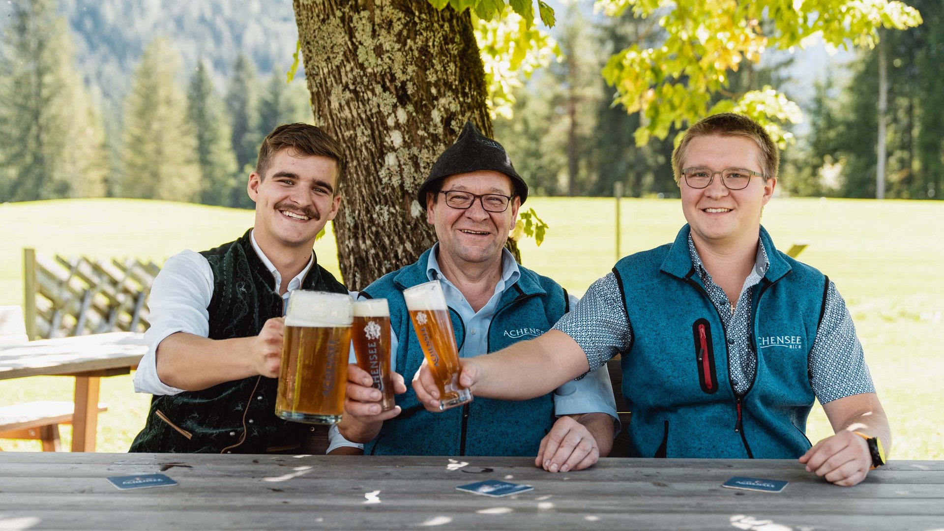 The Karlwirt’s Achenseebier Three men clinking beer glasses outdoors under a tree