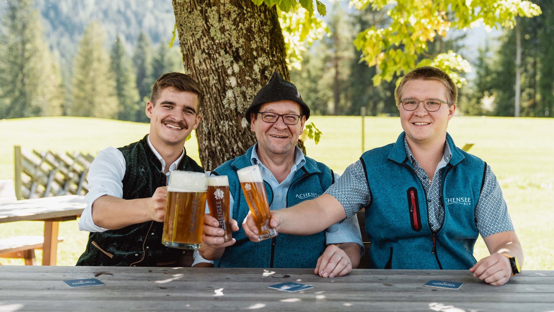 The Karlwirt’s Achenseebier Three men clinking beer glasses outdoors under a tree