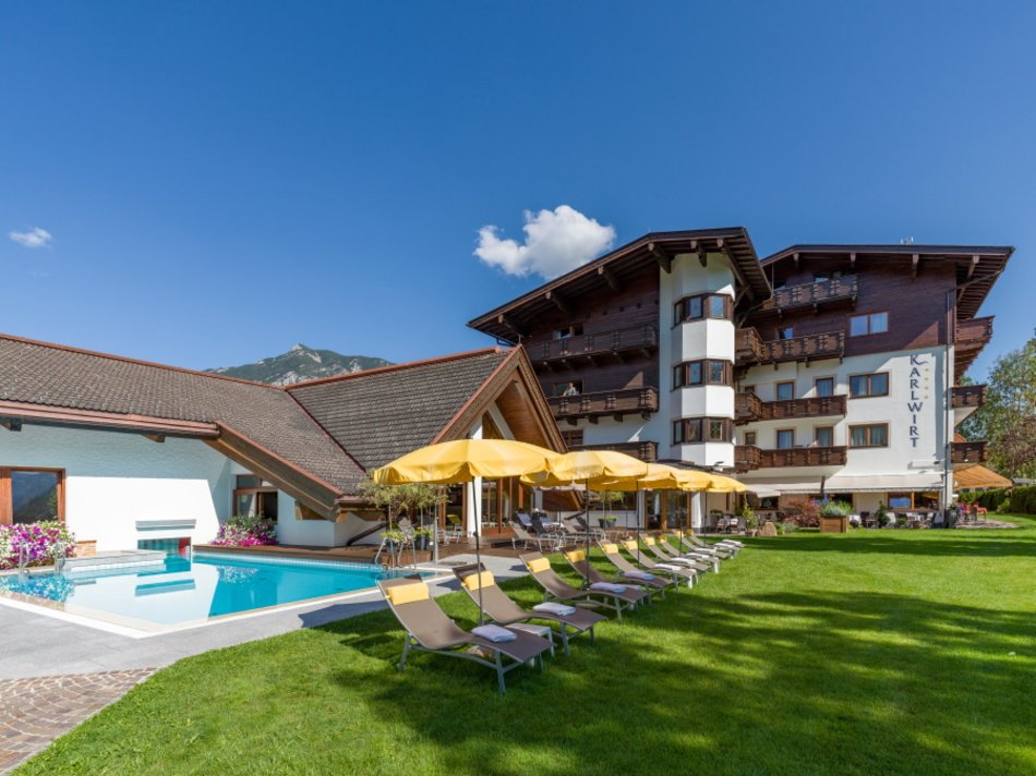 Das Hotel Karlwirt: herzlich willkommen! Hotelgebäude mit Pool, Liegestühlen und Sonnenschirmen auf einer grünen Wiese bei klarem Himmel